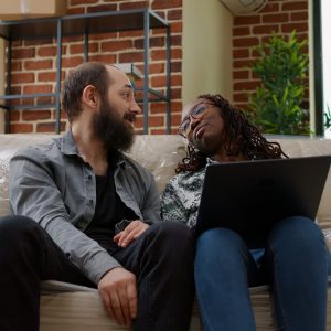 Multiethnic couple finding home decor inspiration on laptop, doing online shopping to move in new rented apartment flat. Unpacking house furniture and belongings for new beginnings.