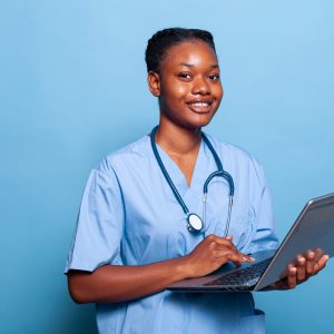 Portrait of african american practitioner nurse smiling at camera while working at medical expertise typing healthcare treatment on laptop computer in studio with blue background. Medicine concept