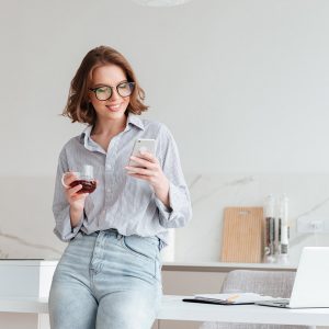 Portrait of a happy attractive woman using mobile phone while holding cup of tea and leaning on a table with laptop computer at home